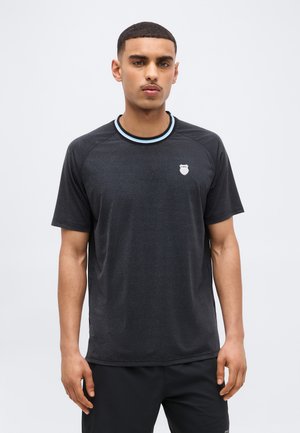 Mężczyzna o krótkich włosach i wąsach nosi czarną koszulę z krótkim rękawem, z białym kołnierzem w paski i małym białym logo na lewej piersi.