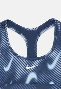 Nike športni modrček z modrim gradientnim vzorcem in navpičnimi črtami. Oblika vključuje stil z odprtinami na hrbtu in belim logotipnim poudarkom.