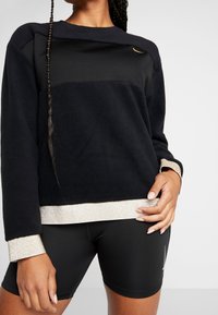 Mustahi sweatshirt mweusi iliyo na mapambo ya dhahabu kwenye mikono na hems, ikiwa na muundo ulioimarishwa na nembo kwenye kifua, ikivaliwa juu ya suruali fupi za mweusi.