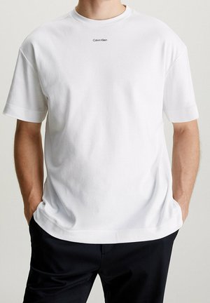 Homme portant un t-shirt Calvin Klein blanc uni à manches courtes, légèrement rentré dans un pantalon noir, les mains dans les poches, sur un fond uni.