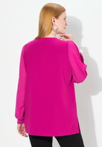 Maglietta fucsia a maniche lunghe con polsini arricciati e spacchi laterali. Tessuto morbido con vestibilità rilassata, vista da dietro.