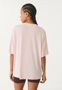 Světle růžové oversized tričko s krátkými rukávy, hladkou texturou, kulatým výstřihem a mírně zakřiveným lemem na zadní straně.