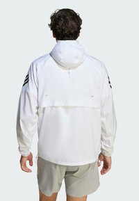 Hvid vindjakke med hætte, der har sorte stribede detaljer på skuldrene og justerbare snore. Glat, letvægtsstof.