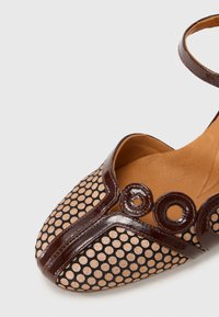 Scarpa in pelle patinata marrone con ritagli circolari, dotata di una tomaia in tessuto beige perforato, dettagli rotondi e cinturino alla caviglia.