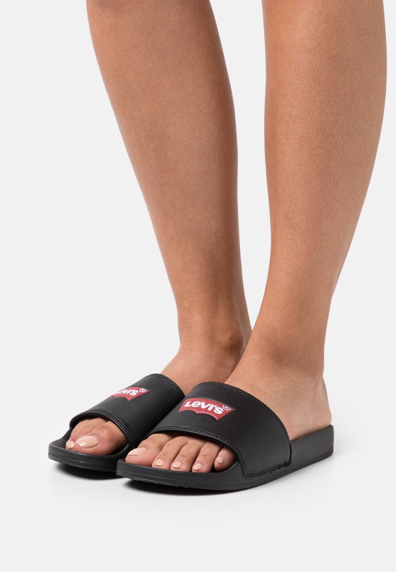 Black slide sandals with wide strap featuring a red Levi's logo. Flat sole, smooth texture, open-toe design, worn on bare feet.