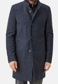 Uomo che indossa un cappotto di lana blu scuro con bottoni neri sopra una giacca nera con zip, in piedi con le mani rilassate lungo i fianchi.