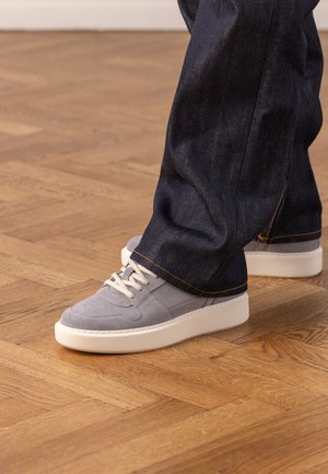 Persoon die donkerblauwe jeans en lichtgrijze suède sneakers met witte veters en dikke witte zolen draagt, staand op een houten parketvloer.
