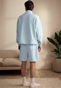 Sweatshirt albastru deschis cu guler înalt și pantaloni scurți asortați. Material moale, croială lejeră și buzunar în spate pe pantaloni. Șosetele nu sunt vizibile.