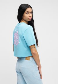 Νεαρή γυναίκα με μακριά σκούρα μαλλιά που φοράει ανοιχτό μπλε κοντό t-shirt με ροζ σχέδιο και ανοιχτό μπλε παντελόνι, γυρνάει να κοιτάξει πάνω από τον ώμο της.