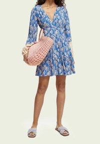 Modré šaty s květinovým vzorem, hlubokým výstřihem do V a dlouhými rukávy. Měkká růžová kabelka a šedé nazouvací sandály dokončují vzhled.