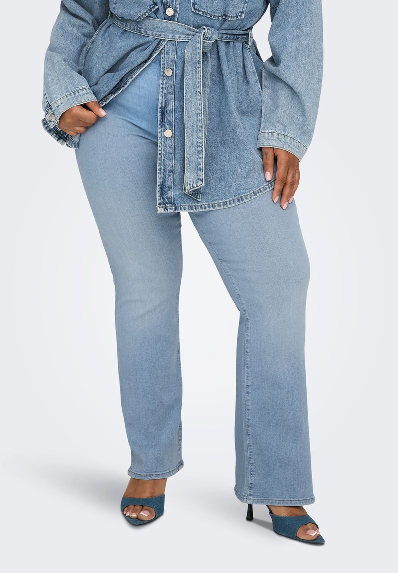 Γυναίκα που φοράει ανοιχτό μπλε τζιν παντελόνι και ταίριαστο τζιν μπουφάν με ζώνη, με μπλε πέδιλα ψηλά τακούνια, σε λευκό φόντο.