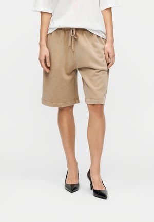 Beżowe szorty z mieszanki bawełny z elastycznym pasem i sznurkiem do wiązania, wyposażone w głębokie boczne kieszenie i luźny krój, zestawione z czarnymi szpilkami.