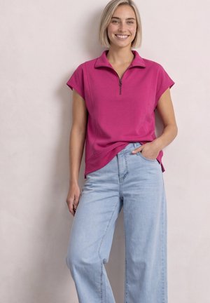 Femme aux cheveux blonds courts sourit, portant un haut rose à manches courtes avec une fermeture éclair et un jean bleu clair à jambes larges, debout devant un fond uni.