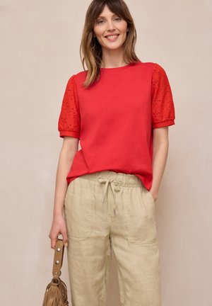 Uśmiechnięta kobieta w czerwonym topie z krótkim rękawem o fakturowanych rękawach i beżowych spodniach ze sznurkiem, trzymająca przy boku brązową torebkę.