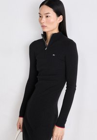 Rochie neagră cu mâneci lungi și dungi, cu guler înalt cu fermoar, având manșete cu accente roșii și un mic logo pe piept. Textură netedă.