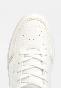 Bout de basket en cuir blanc avec des perforations et des lacets blancs noués sur une languette texturée sur un fond clair.