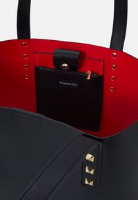 Sac fourre-tout noir avec un intérieur rouge, comportant une petite poche zippée noire et des clous dorés sur le côté extérieur. Fabriqué en matériau texturé.