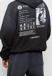 Hoodie noir en tissu doux avec un grand graphique au dos comprenant du texte, des images et des détails d'empreinte digitale. Accompagné d'un pantalon gris.