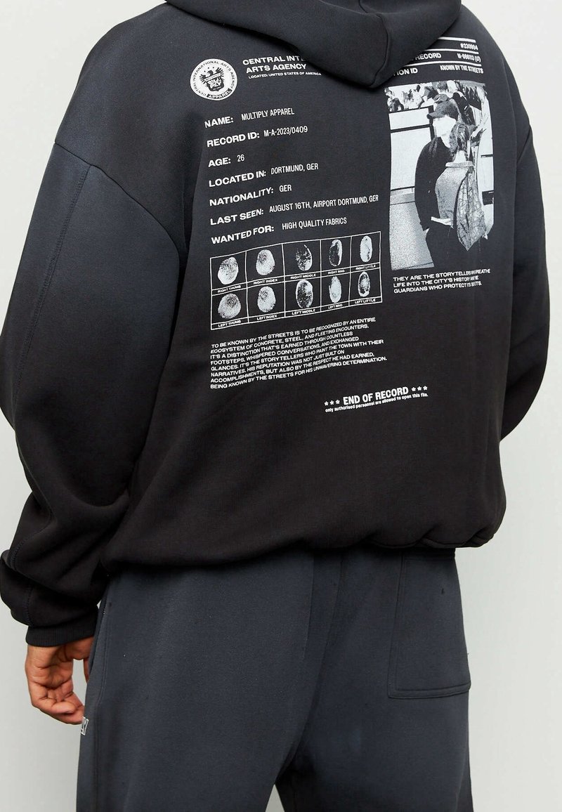 Hoodie noir en tissu doux avec un grand graphique au dos comprenant du texte, des images et des détails d'empreinte digitale. Accompagné d'un pantalon gris.