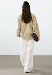 Beige Wildleder oversized Jacke mit einer runden Silhouette, kombiniert mit weißen weiten Hosen. Die Person trägt eine schwarze gewebte Clutch.