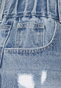 Tela de mezclilla azul claro con un parche desgastado, costuras visibles y un diseño de bolsillo trasero. La textura tiene un aspecto tejido.