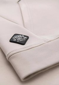 Lys beige sweatshirt stof med en bl�ød tekstur. Har en sort diamantformet lap med prægede bogstaver og logo på ærmet.