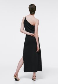 Personne aux cheveux courts portant une robe noire à une épaule avec une fente latérale, debout avec un bras croisé derrière le dos.