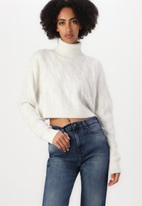 Kremfarget, kort turtleneck genser med kabelstrikkmønster og ripet kant. Kombinert med mørkeblå denimjeans.
