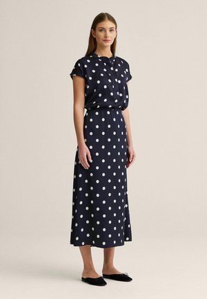 Nainen seisoo beigeää vasten pukeutuneena tummansiniseen, lyhythihaiseen, polkadot-kuvioiseen midimekkoon ja mustiin slip-on-kenkiin.