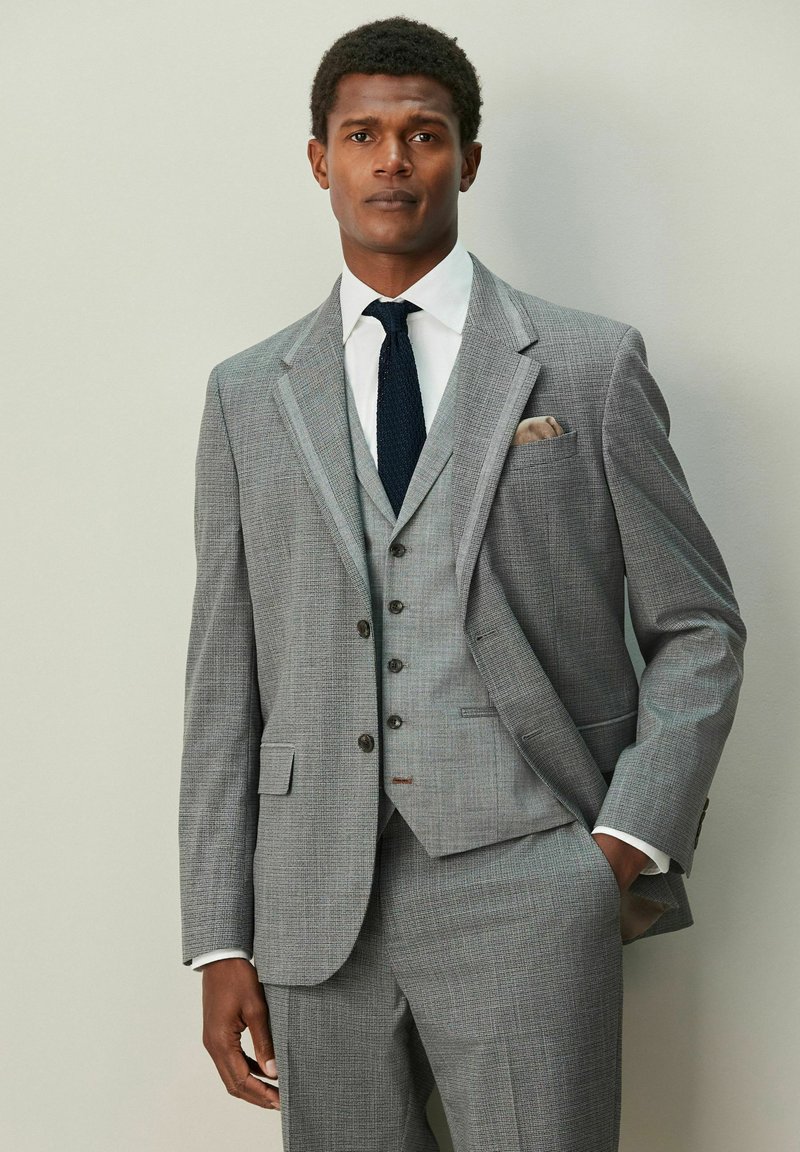 Costum gri din trei piese cu model subtil în carouri. Sacoul are revera cu tăieturi, vestă coordonată și cămașă albă cu cravată neagră.