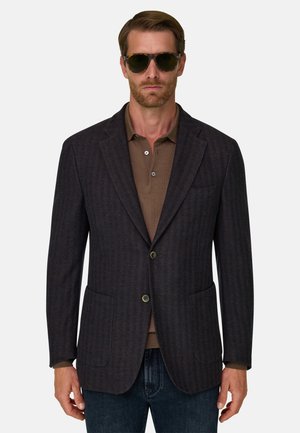 Homme portant un blazer à carreaux foncé sur un polo marron et un jean bleu, avec des lunettes de soleil, debout devant un fond clair uni.