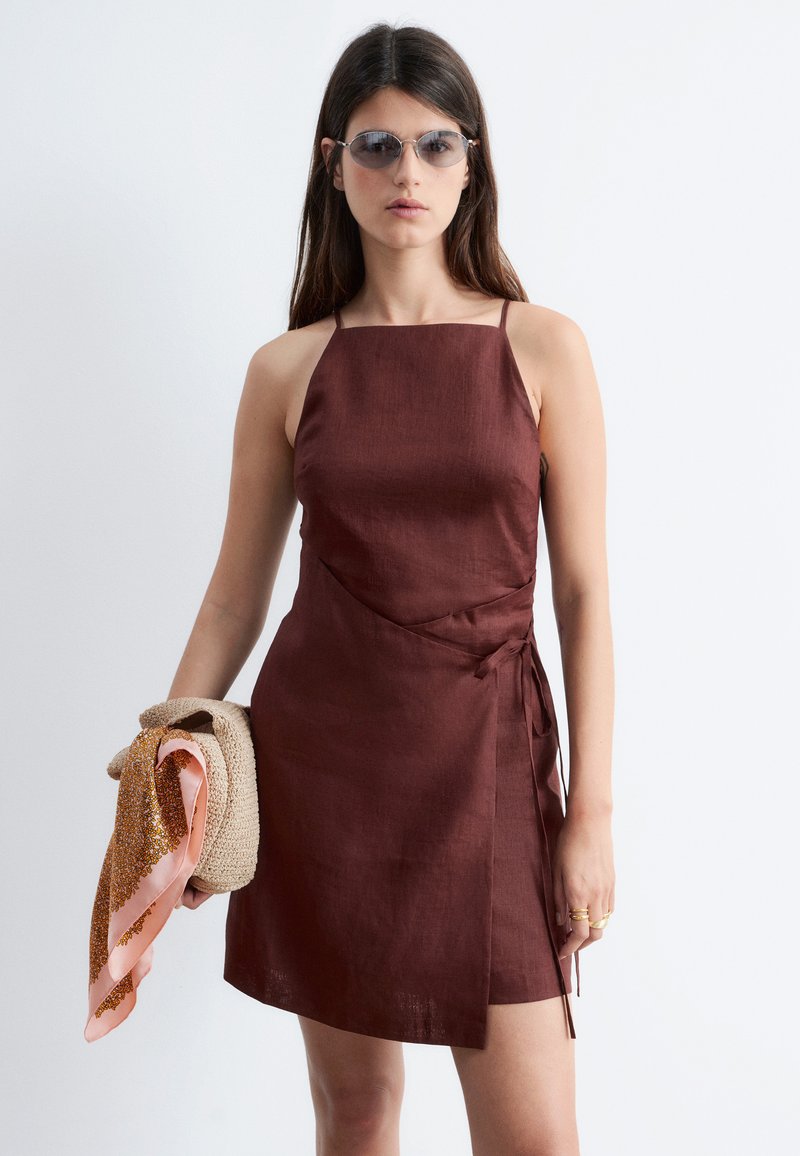Vrouw draagt een mouwloze bruine wikkeljurk en een zonnebril, houdt een beige geweven tas vast met een roze sjaal met patroon.