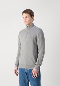 Pull zip gris avec un col montant, des poignets et un ourlet côtelés. Présente un petit logo sur la poitrine, fabriqué en matériel tricoté doux.