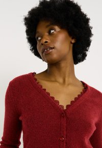 Cárdigan rojo hecho de una tela suave con escote en V y borde de encaje en el cuello. Cuenta con un cierre de botones y mangas largas.