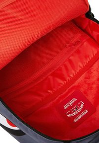Interno di uno zaino in tessuto grigio con fodera rossa brillante, scomparti texturizzati, una tasca con zip e un'etichetta quadrata con i dettagli del marchio.