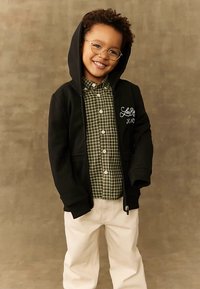 Usmievajúci sa mladý chlapec s okuliarmi, oblečený v čiernej mikine cez zelenú károvanú košeľu a biele nohavice na jednoduchom hnedom pozadí.