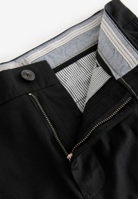 Pantalones negros con cremallera de metal sin cerrar, botón visible y forro interior de la cintura a rayas.