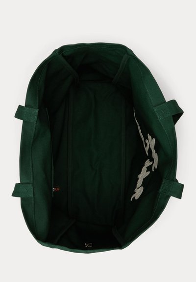 Τσάντα ώμου από καμβά σκούρου πράσινου χρώματος με δύο χερούλια, ανοιχτό πάνω μέρος και εσωτερική πλαϊνή τσέπη, σε θέαση από πάνω σε λευκό φόντο.