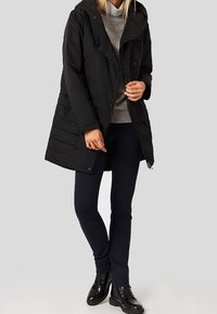 Černý zateplený kabát s kapucí, se zipem a patkami na knoflíky, prošívaný design, nošený přes šedý svetr a tmavě modré kalhoty, doplněný černými botami.