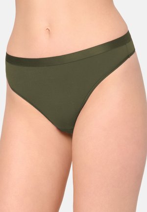 Frau trägt olivgrüne Bikini-Unterwäsche, zeigt die Körpermitte und die oberen Oberschenkel vor neutralem Hintergrund.