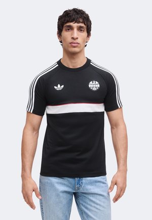 Mand vendt fremad iført en sort Adidas-trøje med hvide striber på ærmerne, et hvidt horisontalt bånd og Eintracht-logo, kombineret med lyseblå jeans.