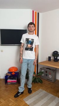 Bílá tričko s grafikou palmy, světle modré džíny, černé tenisky s bílými pruhy, stojící v místnosti s barevnými akcenty.