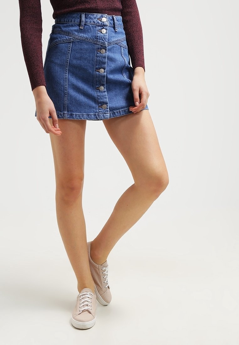 Jupe mini en denim avec fermeture à boutons sur le devant, couleur bleu clair, et un ourlet légèrement évasé. Portée avec des baskets de couleur neutre.