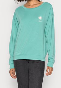 Felpa leggera turchese a maniche lunghe con vestibilità ampia, scollo rotondo e piccolo logo del sole bianco. Tessuto morbido e design semplice.