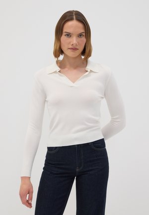 Junge Frau mit schulterlangem braunem Haar, die ein weißes langärmeliges oberteil mit Kragen und dunkelblaue Jeans trägt und vor einem einfarbigen Hintergrund steht.