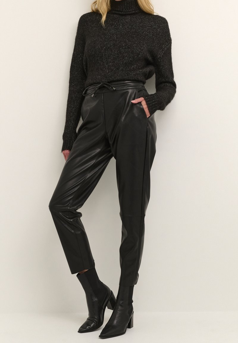 Femme portant un pull à col roulé noir, un pantalon en cuir noir taille haute et des bottines à talons pointues noires.