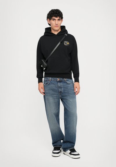 Giovane con capelli ricci e scuri che indossa un felpa nera, una borsa a tracolla, jeans blu larghi e sneakers bianco e nere, in piedi contro uno sfondo bianco.