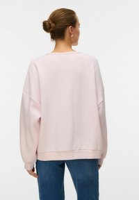 Felpa oversize rosa chiaro con spalle scese e orlo a costine, abbinata a jeans blu, vista da dietro. Morbida texture del tessuto.