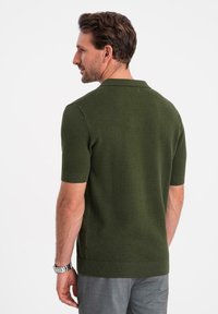 Kortärmad polo i mörkgrön stickad tyg med ett strukturerat mönster, med en krage och ribbade fållar, bärs med grå byxor.