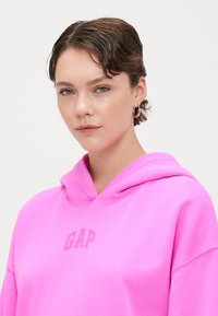 Hanorac cu glugă roz, având logo-ul "GAP" într-o nuanță puțin mai închisă. Textură netedă, croială lejeră și un buzunar frontal tip kangaroo.